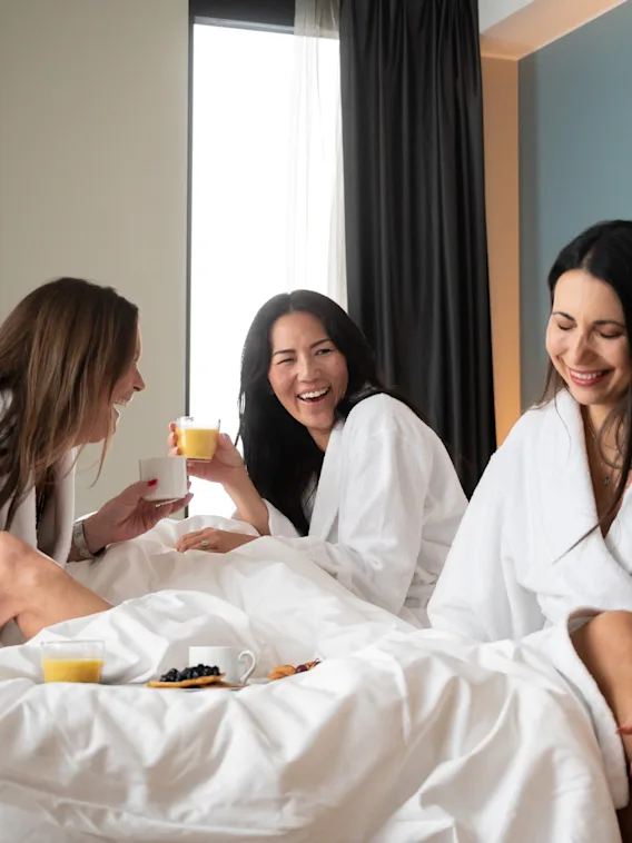Kolme naista valkoisissa kylpytakeissa nauraa ja nauttii juomia sängyllä valoisassa hotellihuoneessa, aamiainen vieressä levitettynä.
