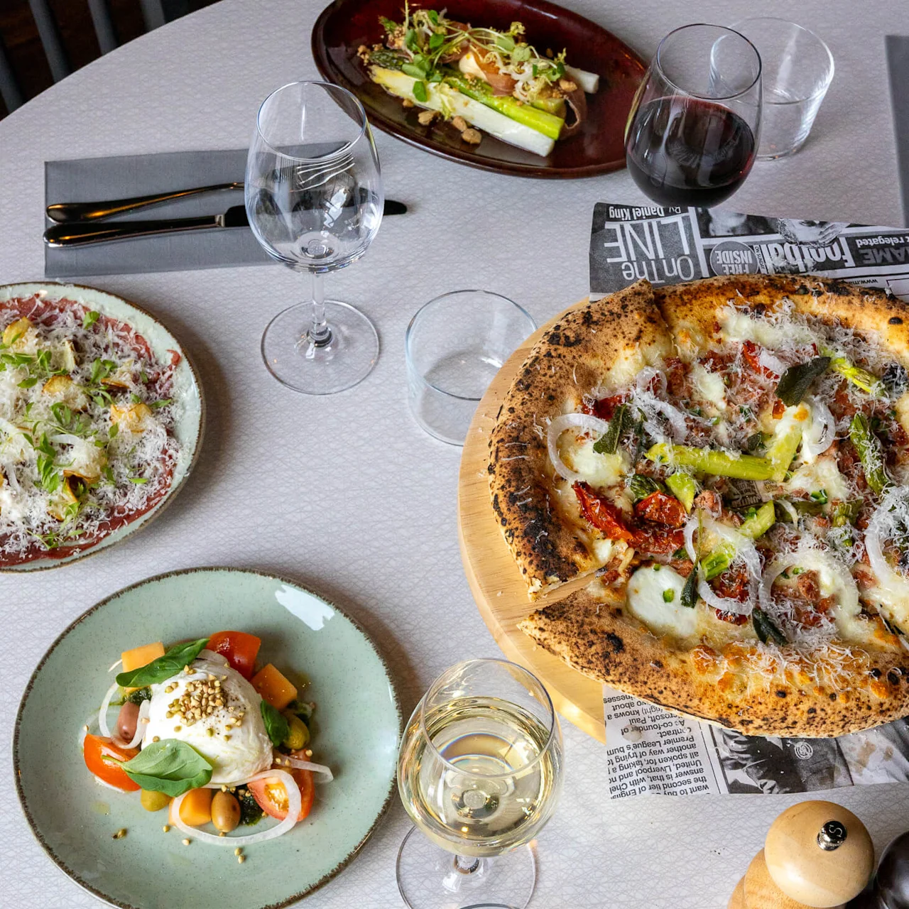 Pizza with onions and herbs sits on a wooden board, surrounded by various salads and wine glasses on a white table, accompanied by cutlery and a pepper grinder.