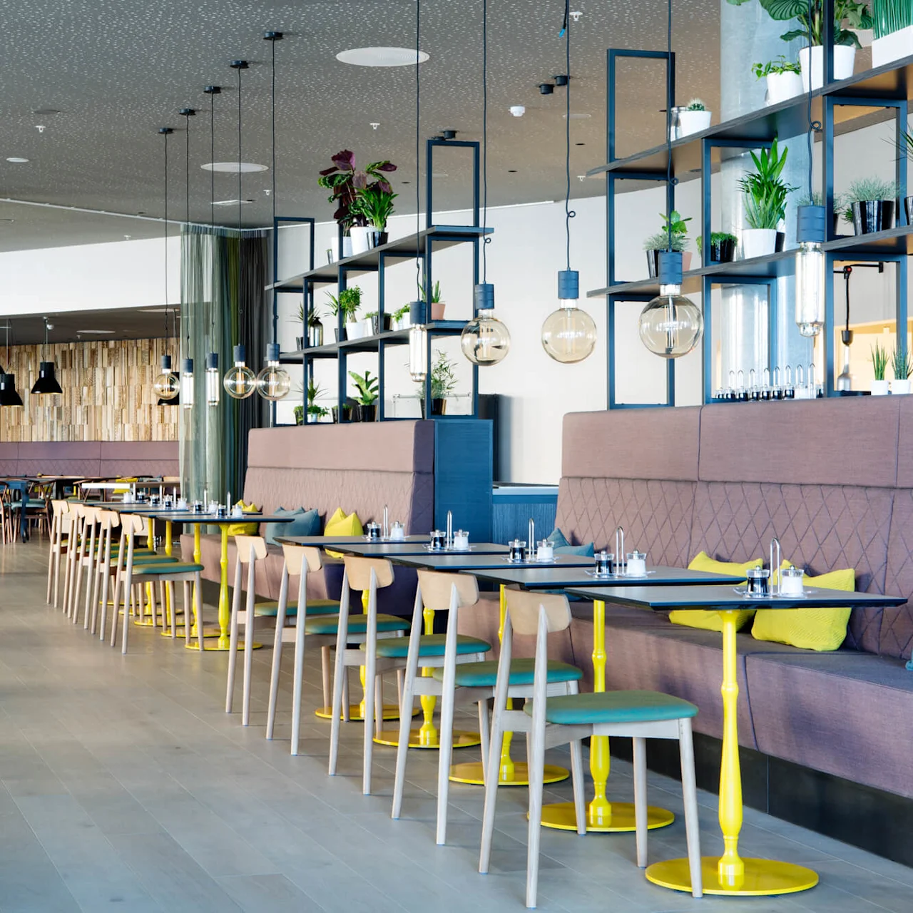Modern dining area with wooden tables, colorful chairs, and vibrant cushions lined along a cushioned bench, under elegant hanging lights. Green plants decorate shelving, creating a lively atmosphere.