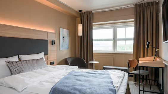 A bed is neatly made with white and blue bedding, situated in a modern hotel room. The room features a desk with a chair, a window with curtains, and wall art.