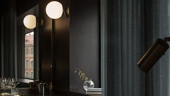 Et svakt opplyst rom har en mørk sofa med puter; en veggmontert globuslampe lyser opp en vinduslist med en vase. Vinglass og dekor på en svart benk.
