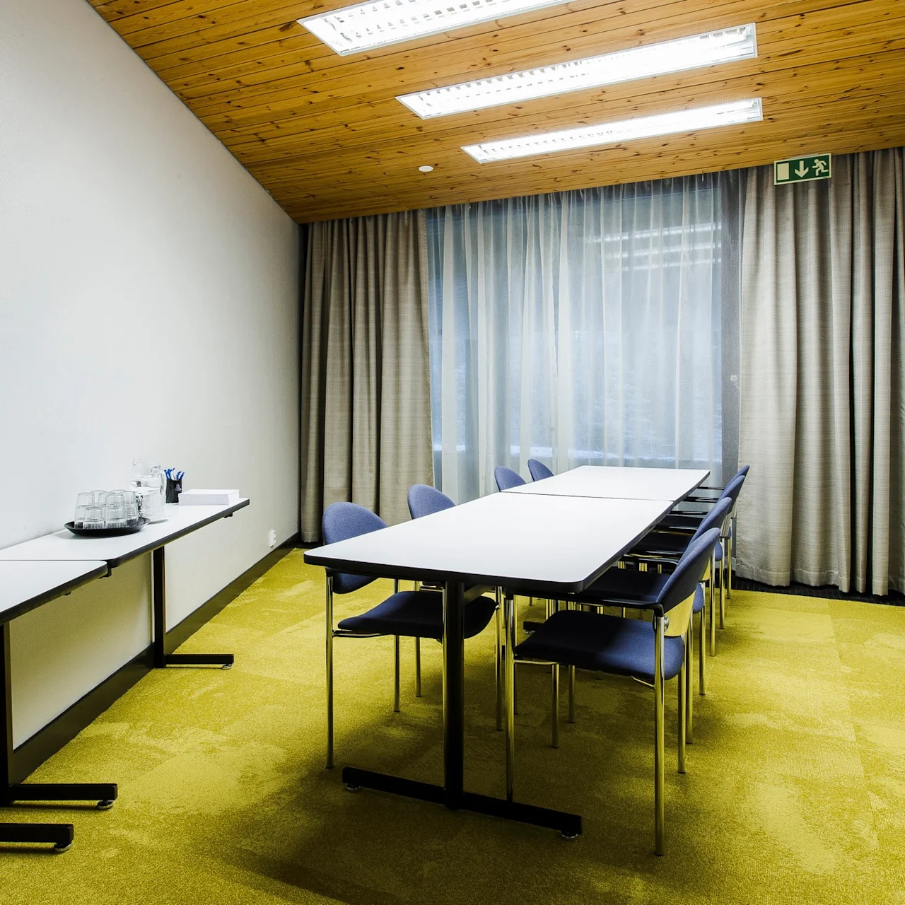 A long, rectangular conference table with several chairs in a room with a wood-paneled ceiling, closed curtains, and a side table holding drinks. An exit sign is overhead.
