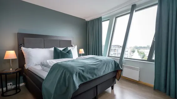 En made dobbeltseng med teal sengetøj og puder står i et moderne soveværelse, flankeret af lamper. Store vinduer afslører en bysilhuet udenfor.