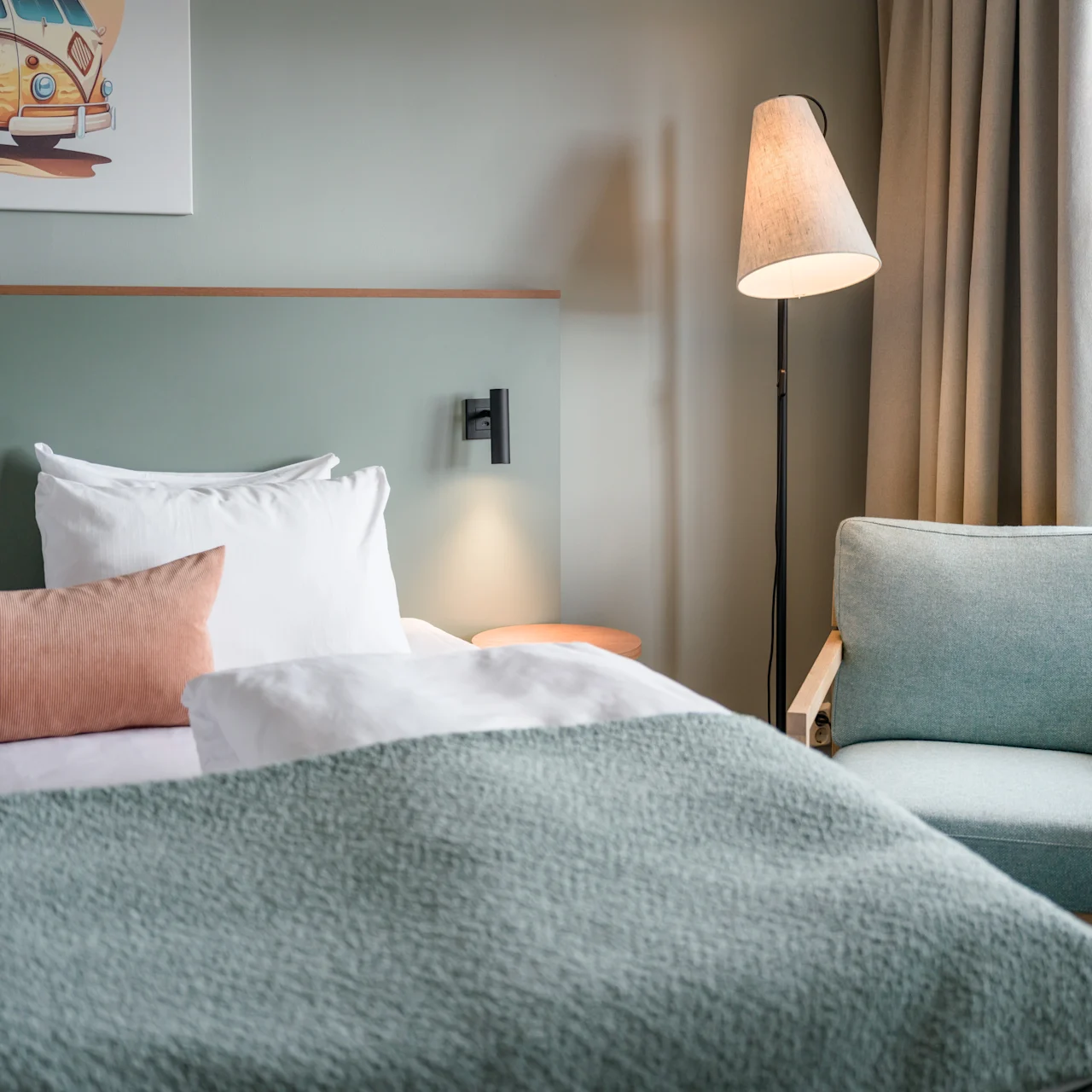 A neatly made bed with white and pastel bedding stands in a cozy room. A painting of a van hangs above; a floor lamp and armchair sit nearby beside a window.