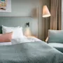 En pent oppredd seng med hvitt og pastellfarget sengetøy står i et koselig rom. Et maleri av en varebil henger over; en gulvlampe og lenestol står ved siden av et vindu.