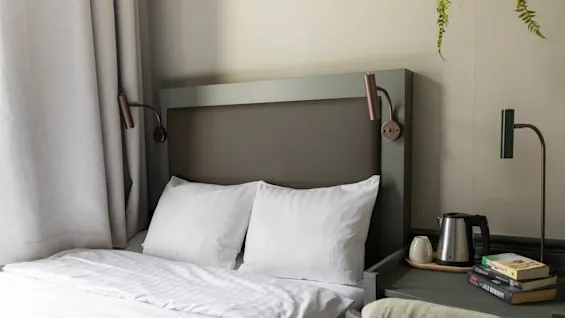 Viihtyisä hotellihuone, jossa siististi pedattu sänky, yöpöytälamput ja pöytä vedenkeittimellä ja kirjoilla, mikä viittaa rentouttavaan oleskeluun.
