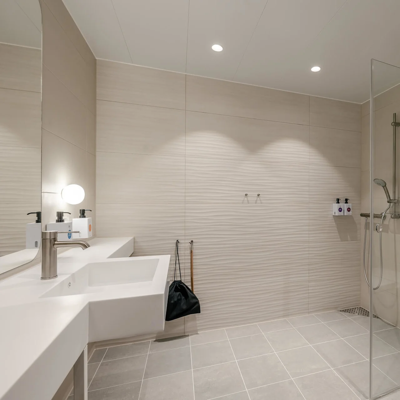 Modern bathroom features a wall-mounted sink and toilet, shower with glass enclosure, tiled walls, and floor. A large mirror and overhead lights enhance the spacious, minimalistic design.