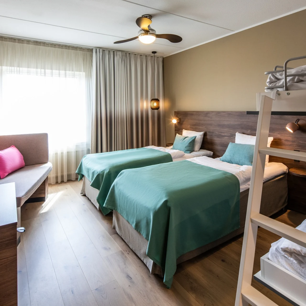 Three beds with green bedspreads are arranged in a neatly furnished bedroom. A ceiling fan spins above, and a sofa with pink cushions sits by a curtained window.