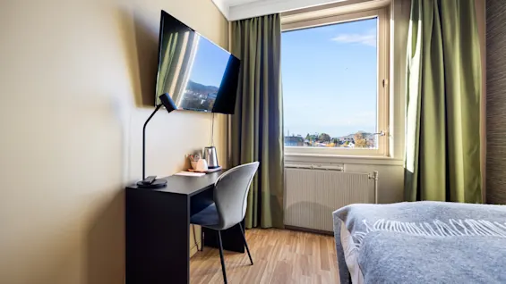 Lyst hotellrom med skrivebord, stol, TV på veggen og et stort vindu som slipper inn dagslys.