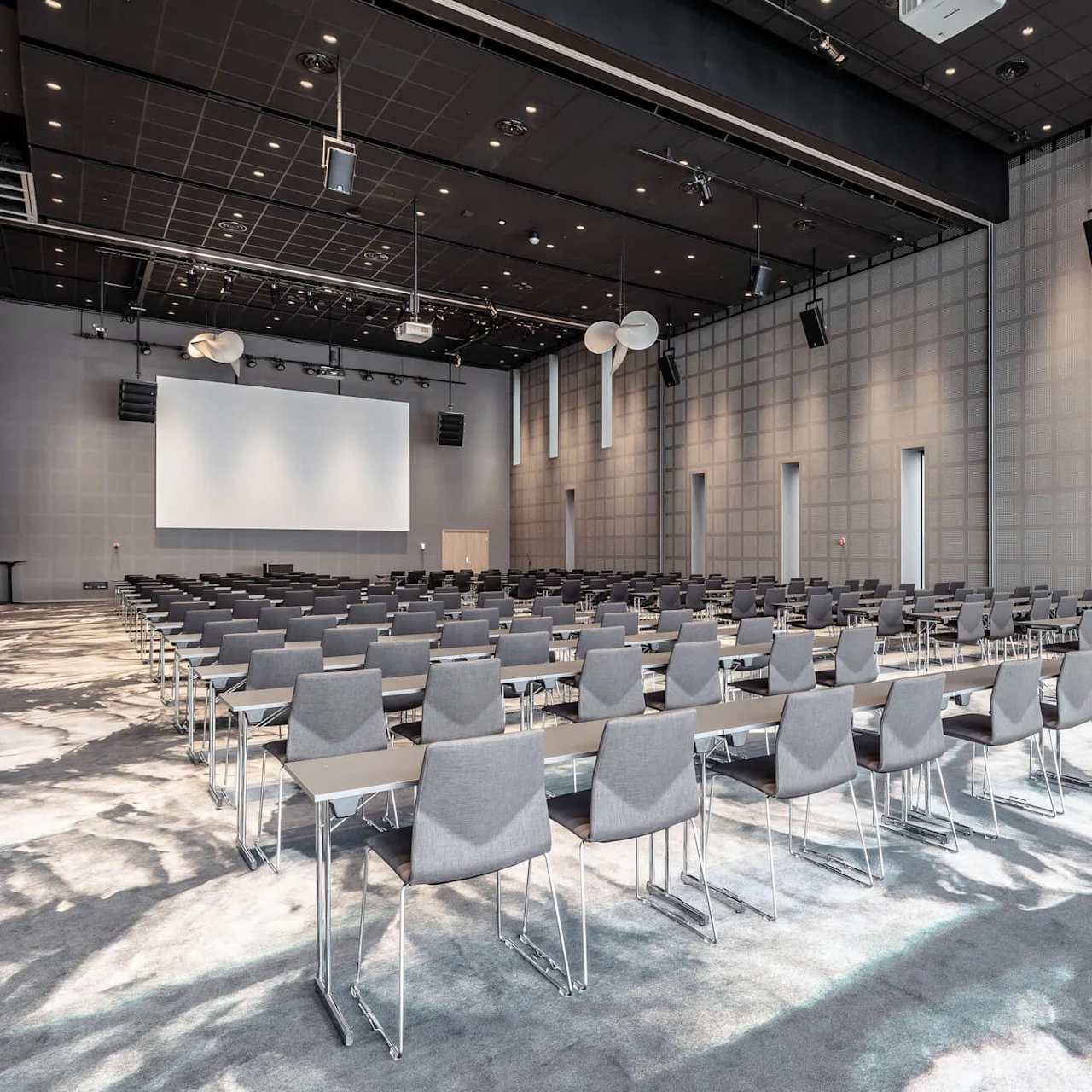 Rows of gray chairs face a large screen in a spacious conference room with high ceilings, soft lighting, and patterned carpet, arranged for a presentation or meeting.