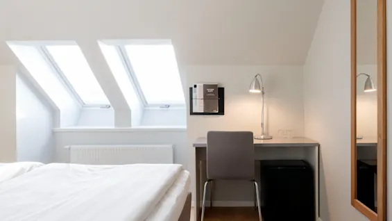 Ett ljust hotellrum med en bekväm säng, ett skrivbord med lampa och två takfönster som ger naturligt ljus.
