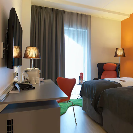 Et moderne hotellrom har to enkeltsenger med fargerike puter, nattbordslamper og et skrivebord med en stol. Et vindu med gardiner gir naturlig lys. En vegg er malt oransje.