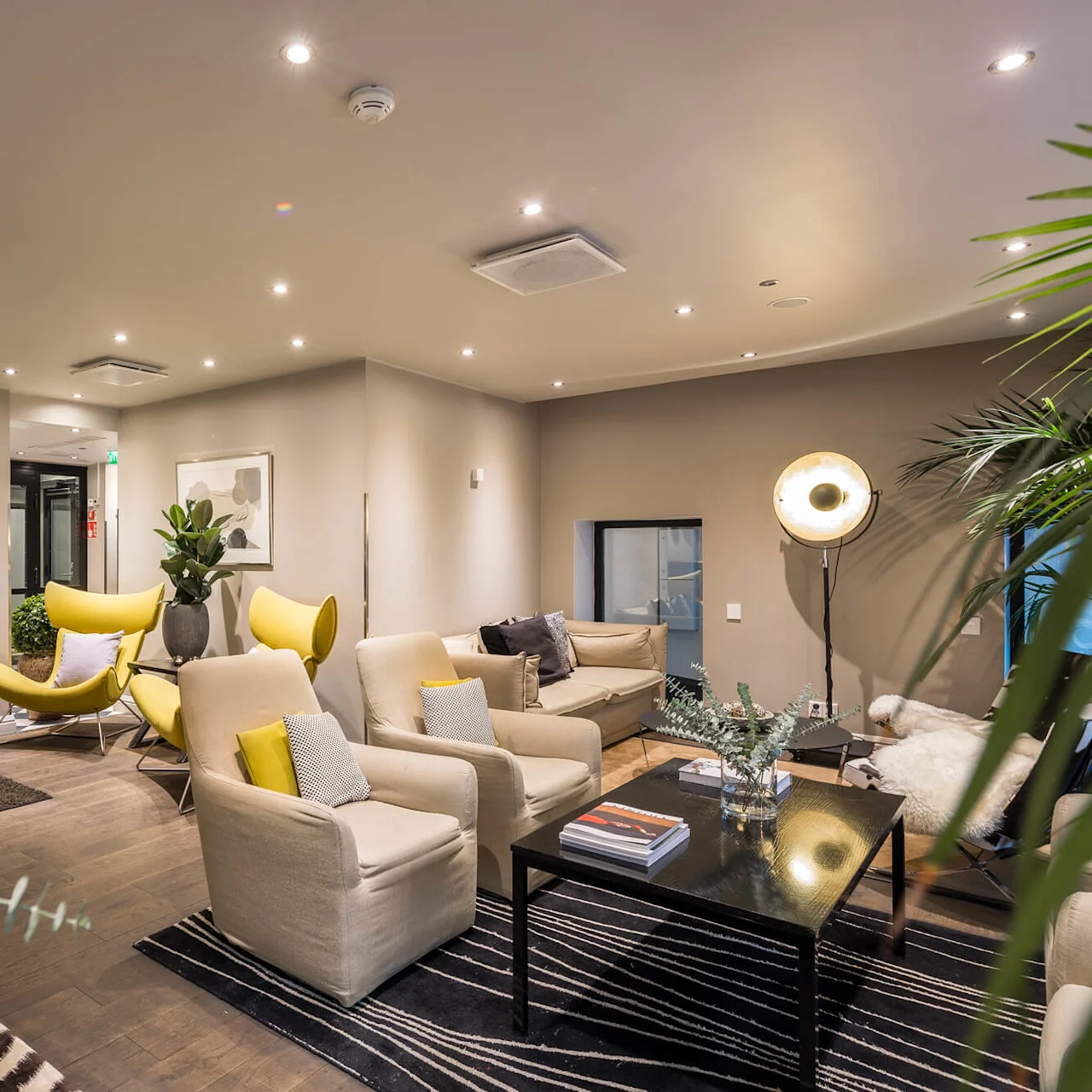Stilig lounge med beige lenestoler og gule stoler rundt et mørkt salongbord, frodige planter, stilfull belysning i en moderne setting med tregulv og subtil dekor.