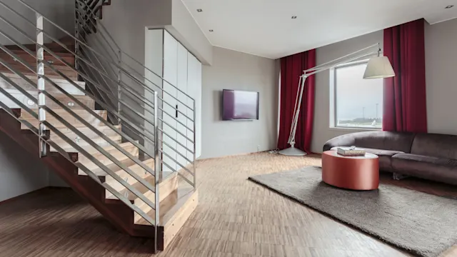 Trapp ned mot en moderne stue med en stor gulvlampe. Rommet har en buet sofa, et rundt bord og en veggmontert TV, vinduer med røde gardiner.