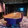 A wooden conference table surrounded by purple chairs stands on a patterned carpet in a modern room with a TV on a dark wall, framed by curtains and artwork.