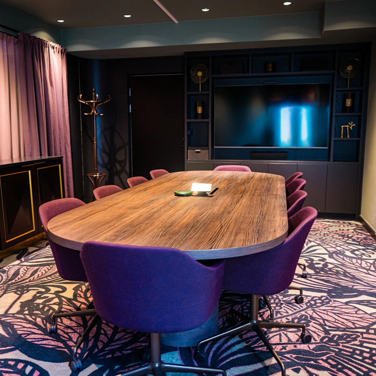 A wooden conference table surrounded by purple chairs stands on a patterned carpet in a modern room with a TV on a dark wall, framed by curtains and artwork.