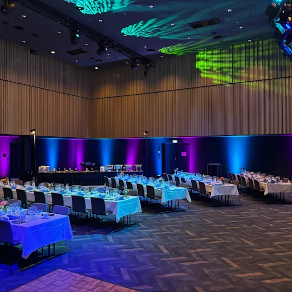 Suuri konferenssihuone, jossa on pöytiä ja tuoleja tapahtumaa varten, värikkäällä valaistuksella. Konferenssi Clarion Hotel Helsinki Airportissa.