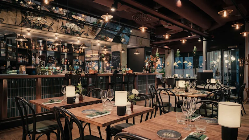 Restaurant tables are set with plates, glasses, and napkins under warm lighting; the background features a well-stocked bar with various bottles and a mirrored wall reflecting the cozy atmosphere.
