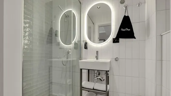 Ett modernt badrum med upplysta speglar och en glasdusch, vita kakel, handfat, handdukar och en svart "Hair Dryer" -väska som hänger på väggen.
