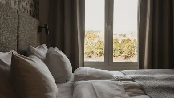 Ett mysigt hotellrum med en prydligt bäddad säng, mjuka kuddar och ett fönster med utsikt över gröna träd och avlägsna byggnader.