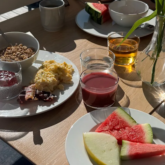 En herlig hotellfrokost i solen med eggerøre, frisk melon, granola og juice på et trebord.