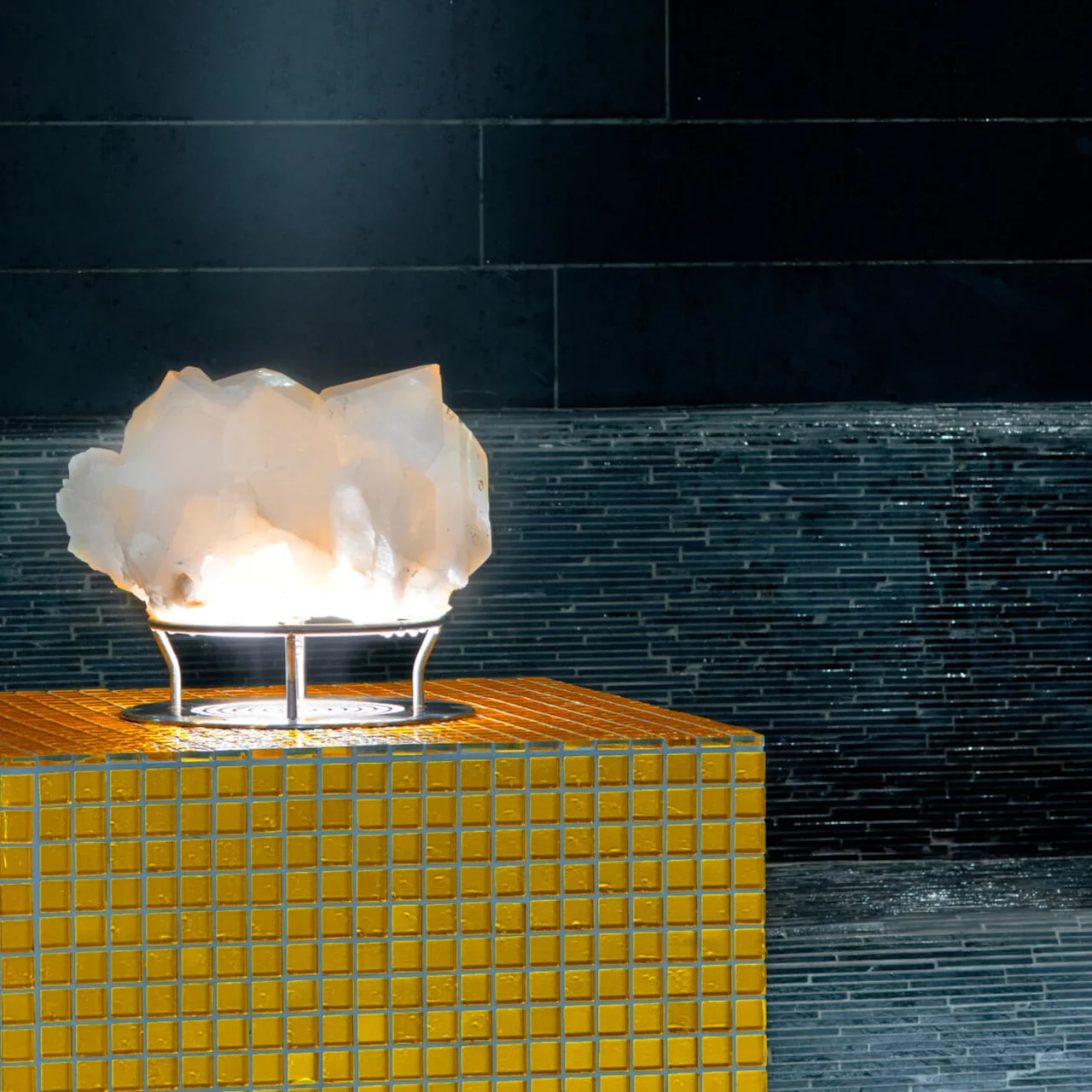 En glødende hvit krystall hviler på et opplyst metallstativ, oppå en gul-flislagt sokkel, mot en mørk, flislagt vegg i et elegant, minimalistisk rom.