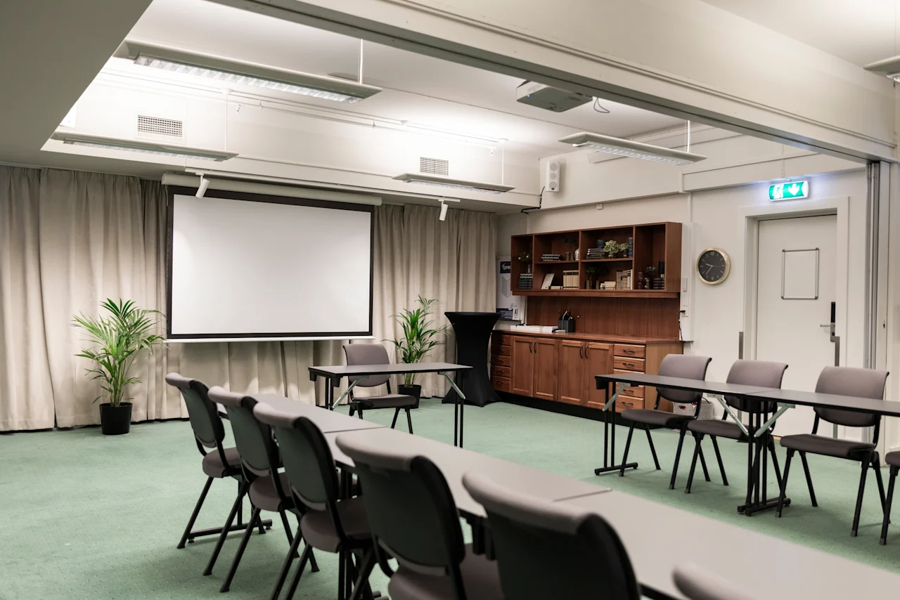 A functional meeting room at Home Hotel Grand Bodø with tables, chairs, a projector screen, and an EXIT sign. Ready for presentations.
