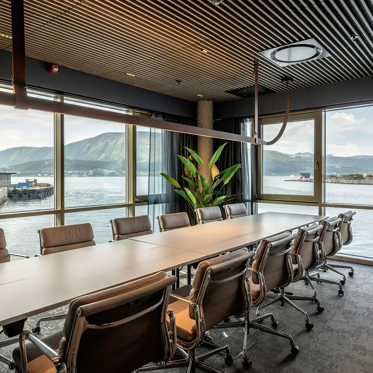 Konferencebord omgivet af grå stole, sat mod store vinduer, der giver udsigt over en naturskøn vandfront og bjerge. Indendørs plante ved vinduet, træloft med moderne belysning.