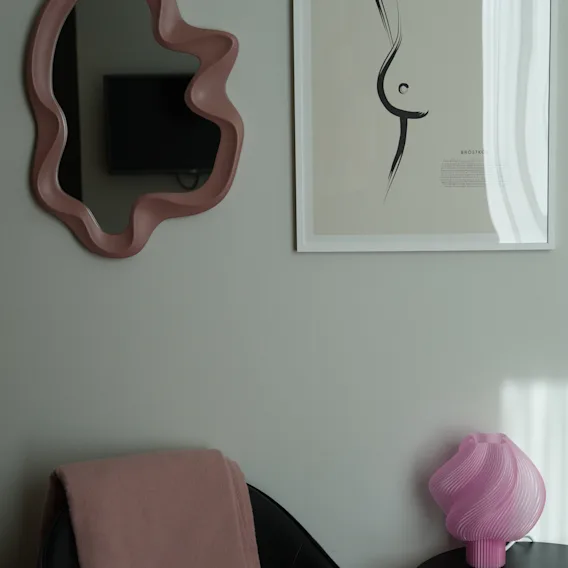 Stilrent hotelværelse med et lyserødt bølget spejl, minimalistisk kunst, en lænestol med tæppe og en lyserød bordlampe.