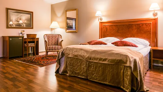A bed with two red pillows sits in a warmly lit hotel room. Nearby, a striped chair and a small desk are positioned on a patterned rug.