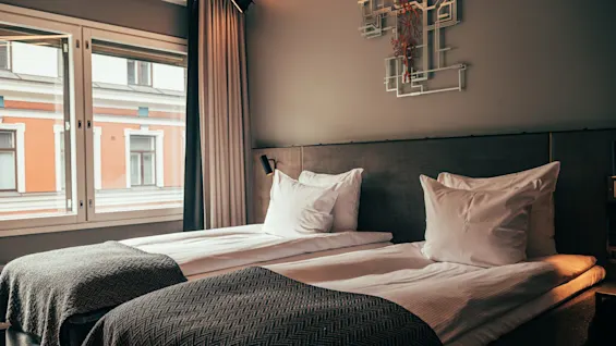 A modern hotel room with two single beds, white bedding, and dark throws, next to a window overlooking a building.