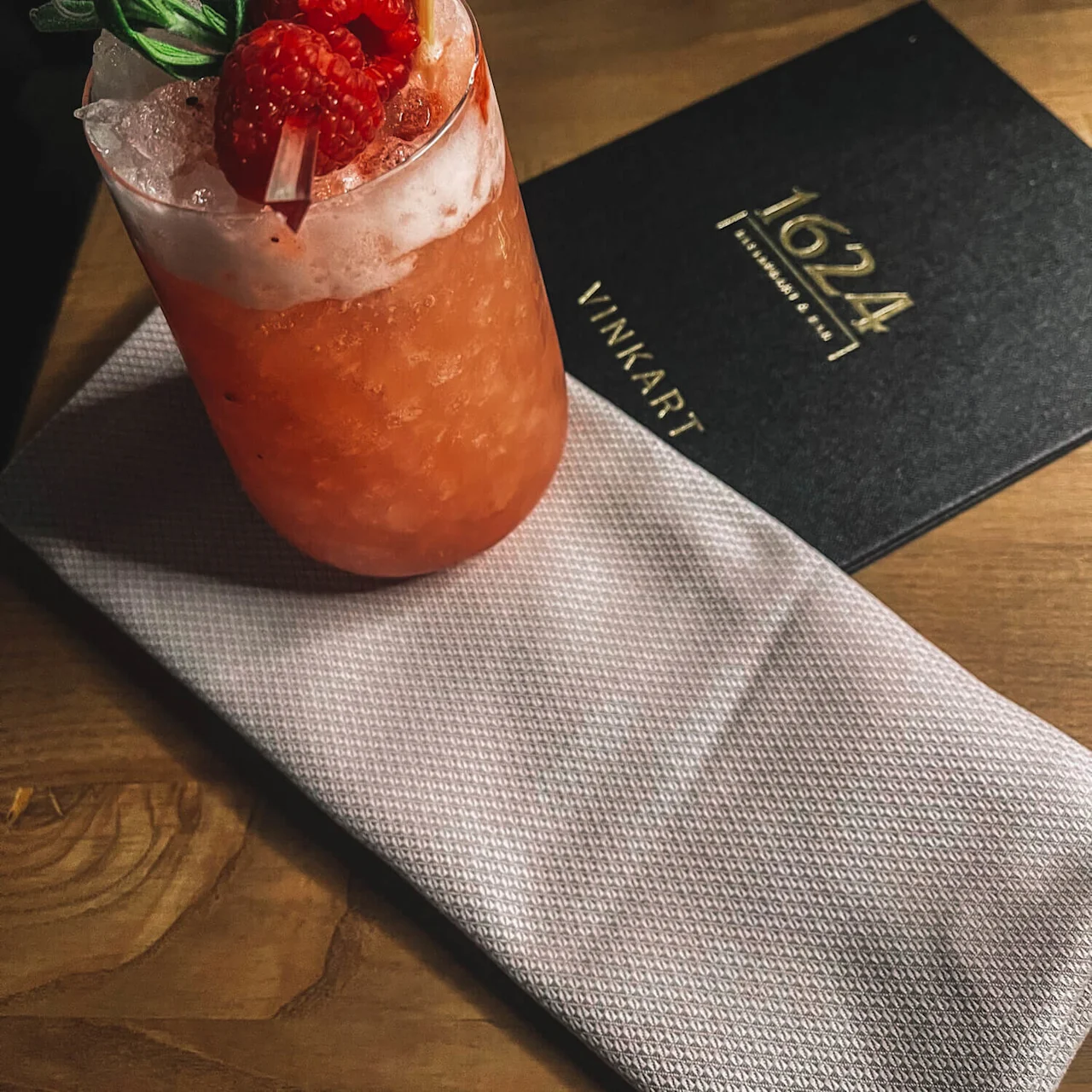A cocktail with raspberries and a sprig of rosemary on top, placed on a mat with a menu reading "1024 VINNART".