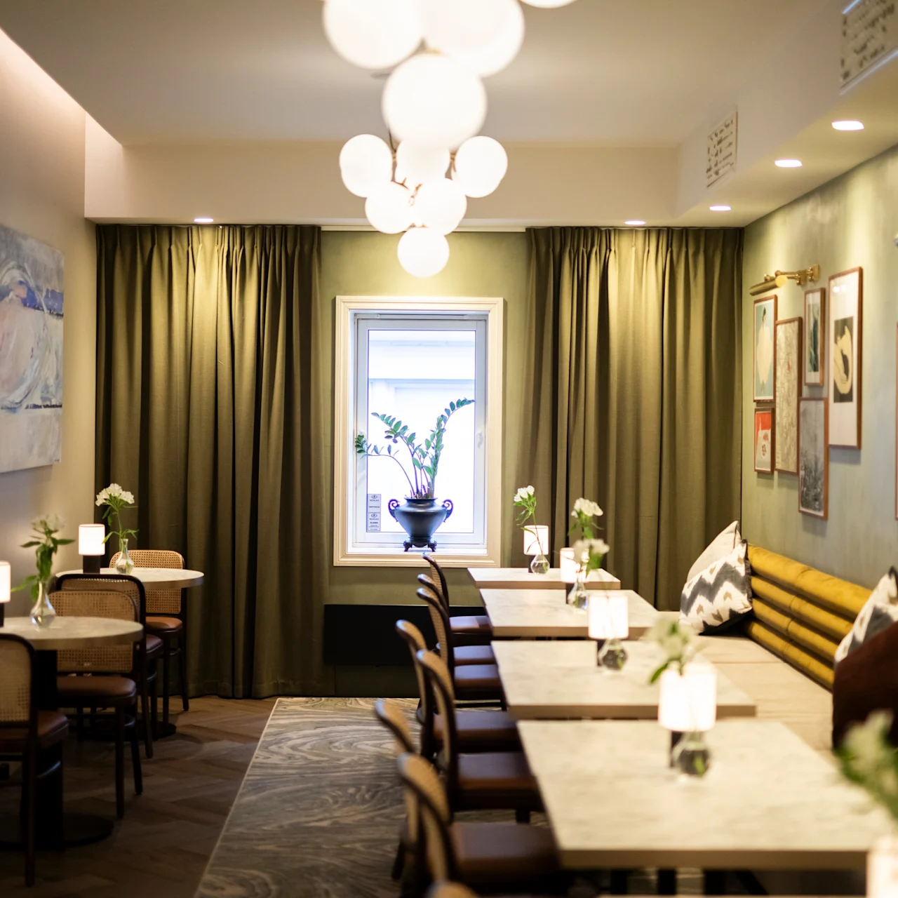A row of marble tables with chairs and decorative plants lies in a cozy, softly lit café. Wall decor and curtains frame the scene, adding warmth and sophistication.