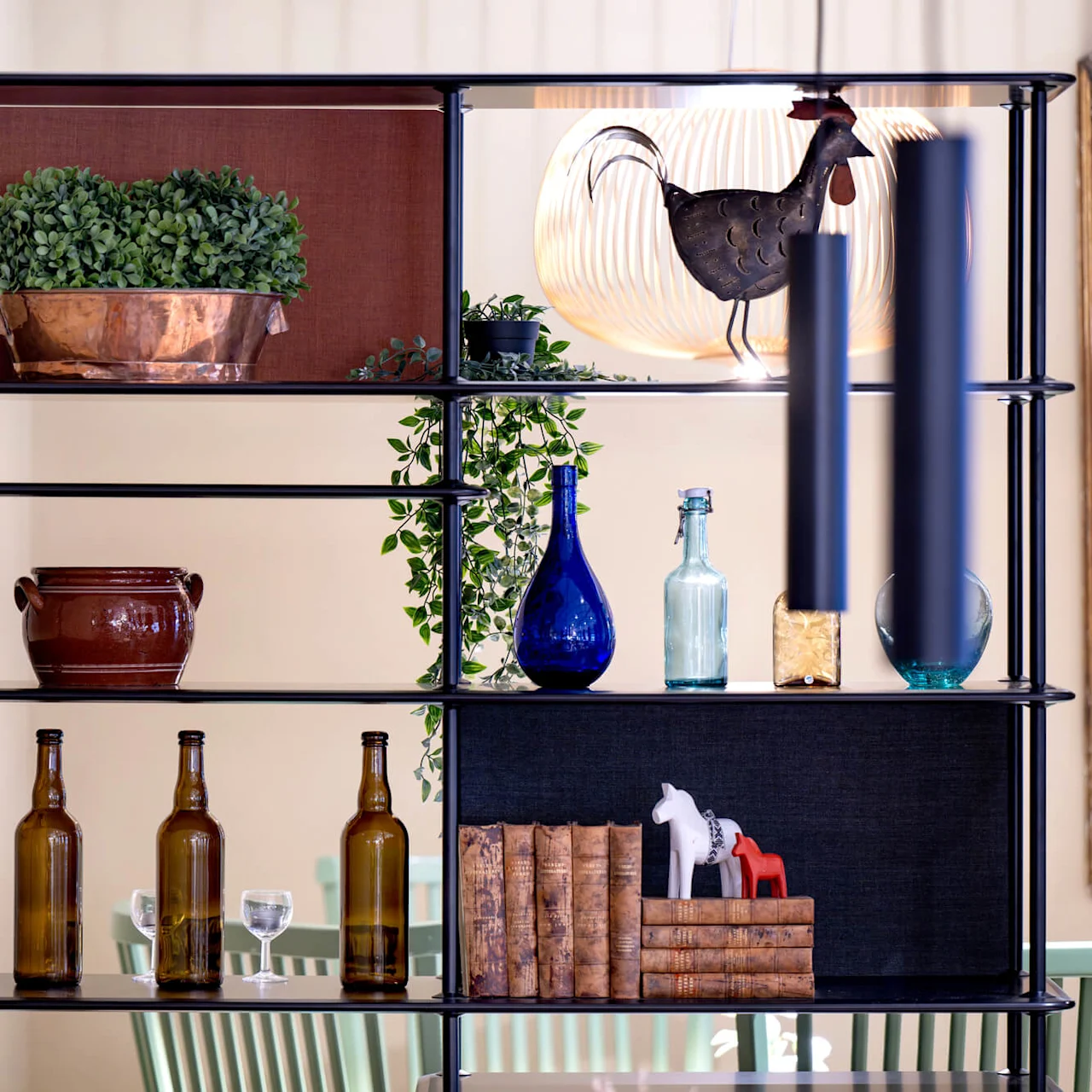 A bookshelf with decorative objects, including plants, bottles, books, and a metal rooster sculpture, set against a warm interior.