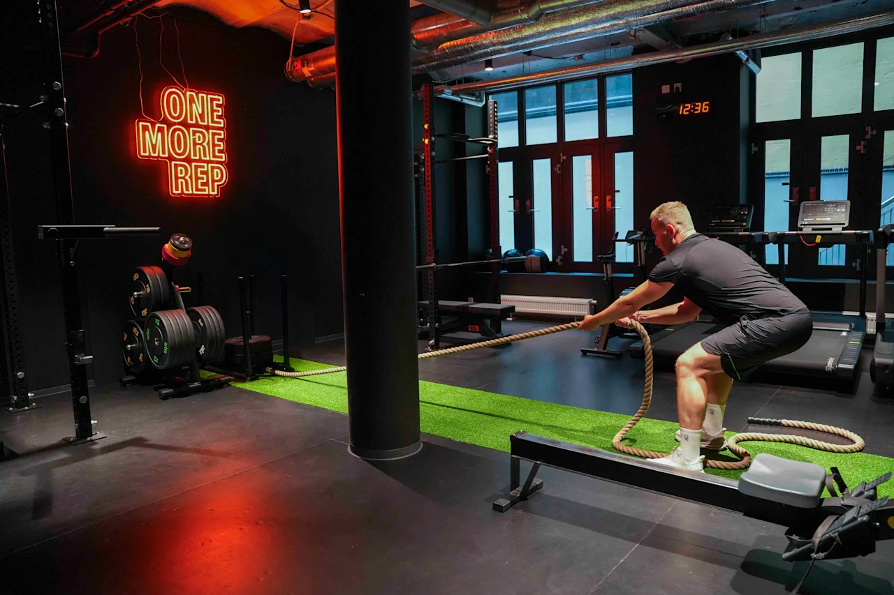 En person trækker et tungt reb indendørs, omgivet af træningsudstyr; neon skiltet siger "ONE MORE REP" og væguret viser 12:36.