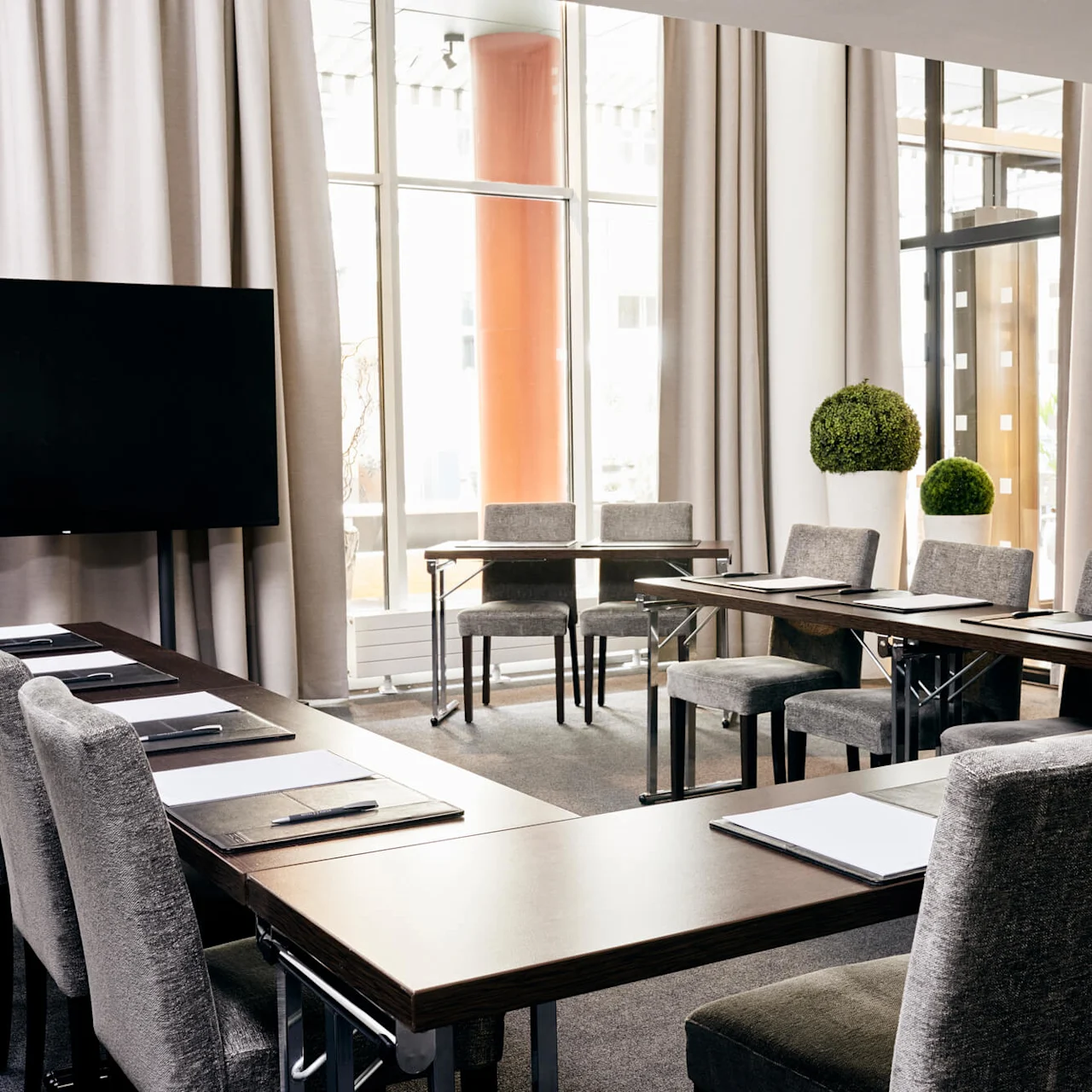 A modern conference room with U-shaped tables and grey chairs, featuring a black television, notepads, and pens. Large windows provide natural light, framed by light curtains.