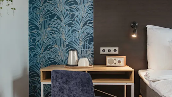 Desk with blue chair and metallic kettle under a lamp, beside a bed with wooden headboard, against a patterned blue wallpaper. Wall outlets and radio nearby.