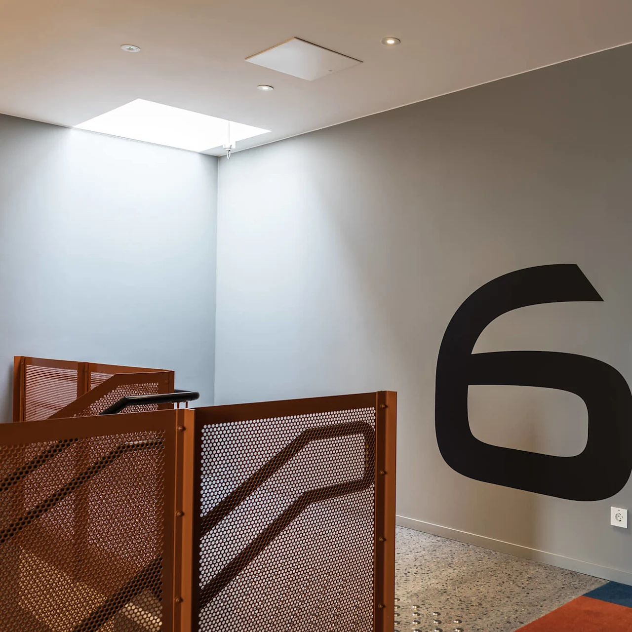 Large black number "6" on a gray wall. Brown railing beside stairs illuminated by overhead light. Adjacent open door leads to corridor with room numbers "601-605" and "606".