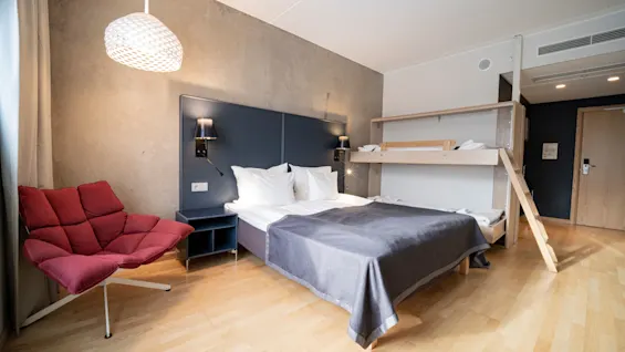 Bed with gray blanket against a wall, beneath a bunk bed. Red chair nearby on wooden floor. Lit wall lamps and a white pendant light hang above.