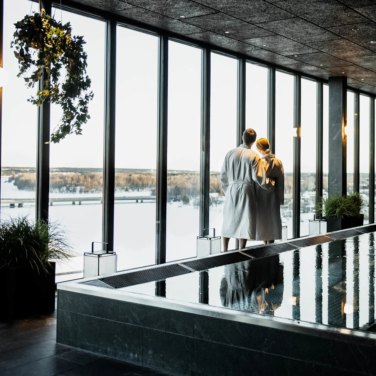 Et par i hvide badekåber står ved store vinduer og kigger ud på et snedækket landskab. De er i et moderne spa-rum med en reflekterende pool og loungestole.