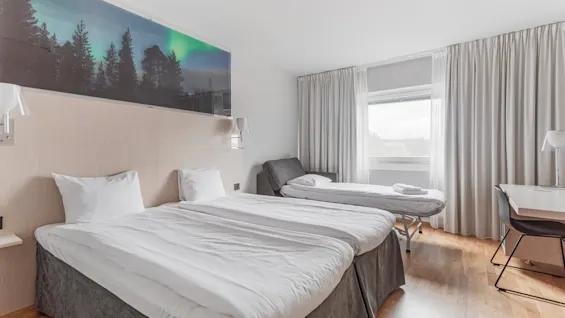 A bright hotel room with two single beds, a rollaway bed, a desk, and a mural of a forest with the Northern Lights.