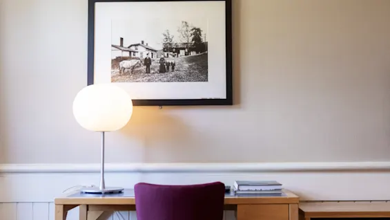 Skrivebord med en lysende lampe står mod en beige væg; ovenfor hænger et indrammet sort-hvidt fotografi af personer foran bygninger. En lilla stol er placeret ved skrivebordet.