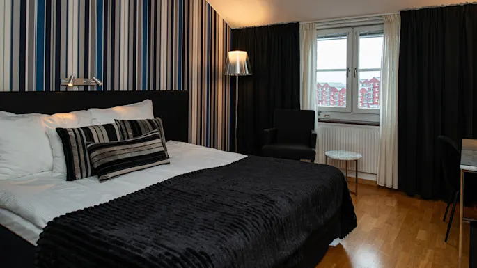 En pænt redt seng med sort-hvide stribede puder og et mørkt tæppe står i et moderne soveværelse. En stol og et lille bord står ved et vindue, med stribet tapet.