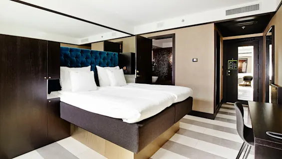 Seng placeret centralt, med hvidt sengetøj og puder, mod en blå tufted sengegavl i et moderne, minimalistisk indrettet hotelværelse med cremefarvede vægge og stribede gulve.