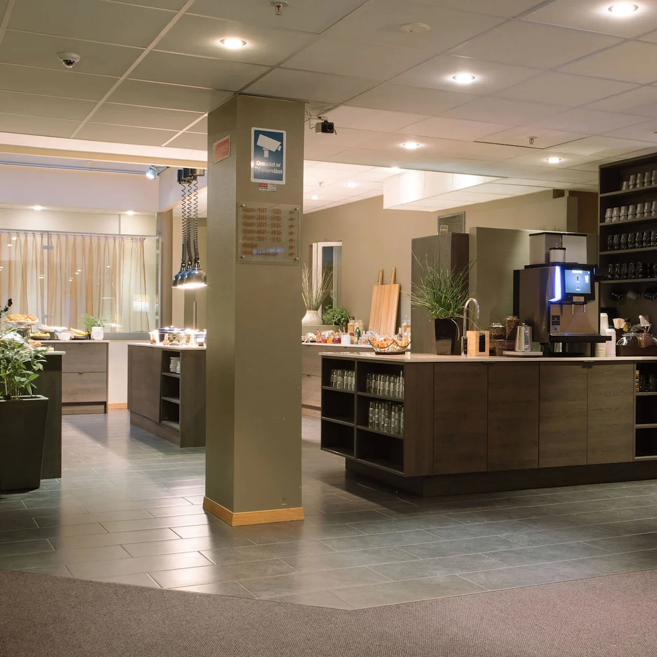 Kahviasema hotellin aulassa, jossa kahvikone, pinotut kupit ja välipalat, ympärillä puiset kaapit ja viherkasveja lämpimässä valaistuksessa. Teksti: "COMFORT HOTEL PARK".