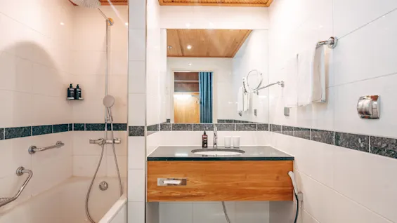 A modern bathroom with a bathtub and shower head, featuring a wooden vanity with a mirror, surrounded by white tiles and ambient lighting. Two bottles are on a shelf.