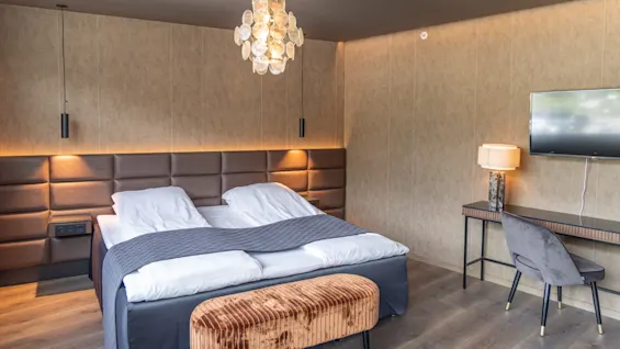 Ett modernt hotellrum med en dubbelsäng med vita sängkläder och grå filt, omgiven av hängande lampor. En sammetspall står vid fotändan. Skrivbord med stol och lampa, TV monterad på väggen.