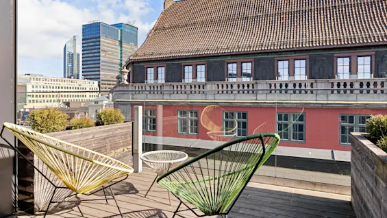 Two colorful chairs sit on a wooden deck, bordered by planters, overlooking a historic building with a contemporary cityscape in the background.