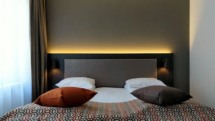 Siististi pedattu sänky, jossa on valkoiset tyynyt ja värikkäät koristetyynyt, on keskitetty tumman sängynpäädyn eteen, huoneessa, jossa on minimalistinen sisustus.