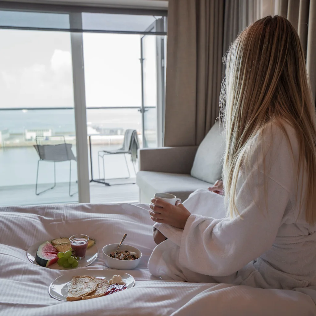 En person i morgenkåpe nyter frokost på sengen med utsikt over vannet fra et hotellrom.